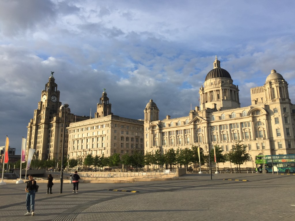 The 3 ornate buildings known as The Three Graces in Liverpool. The Royal Liver Building is the tallest of the three, and has a tall spire with clock faces on each side, and a bird with outstretched wings on the top. The Cunard Building is shorter, and is a very simple rectangular box shape, but it's still very wide and deep, The Port Of Liverpool building is the widest, with a small domed spire on each corner, and a large dome on top of a raised section in the middle.