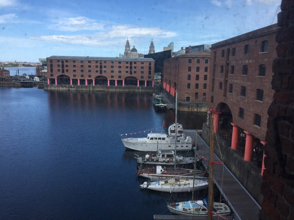 Royal Albert Dock. The large square expanse of water is surrounded by wide brick buildings on each side. Each building is 5 stories high, with large archways and red pillars at the bottom. The River Mersey is visible through an opening between buildings at the top left, and another exit from the dock is mid-way between two buildings on the right.