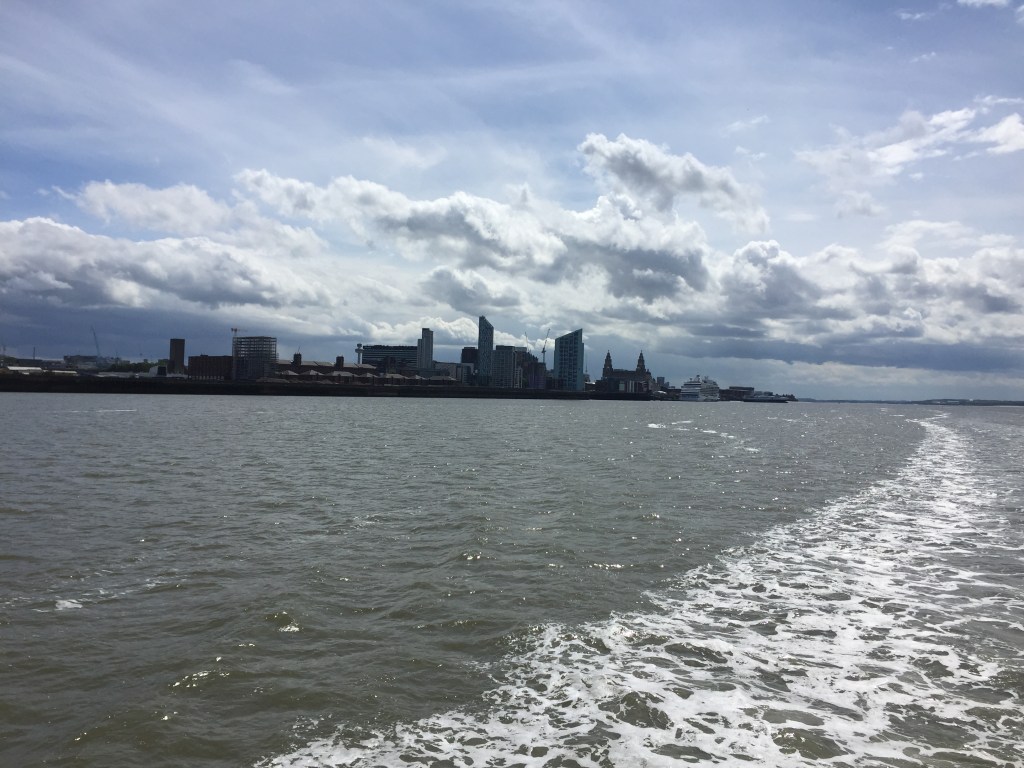 View from the Mersey Ferry towards Liverpool. The front and rear spires of the Royal Liver Building can be seen in the distance, along with a large cruise shop moored near it. A curved trail of frothing water, left behind by the cruise during its journey, is also visible.