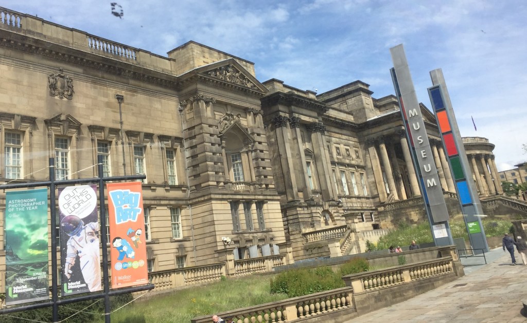 The large World Museum building in Liverpool, the entrance of which is under a high canopy held up by tall pillars. Posters outside the museum advertise the Astronomy Photographer Of The Year, The Planetarium and Big Art For Little Artists.