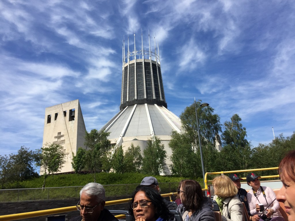 Liverpool Metropolitan Cathedral. The large building resembles an upturned cone with a wide circular tower on top. The top third of the tower is open with tall vertical metal poles around the circumference, linked in between by metal railings.