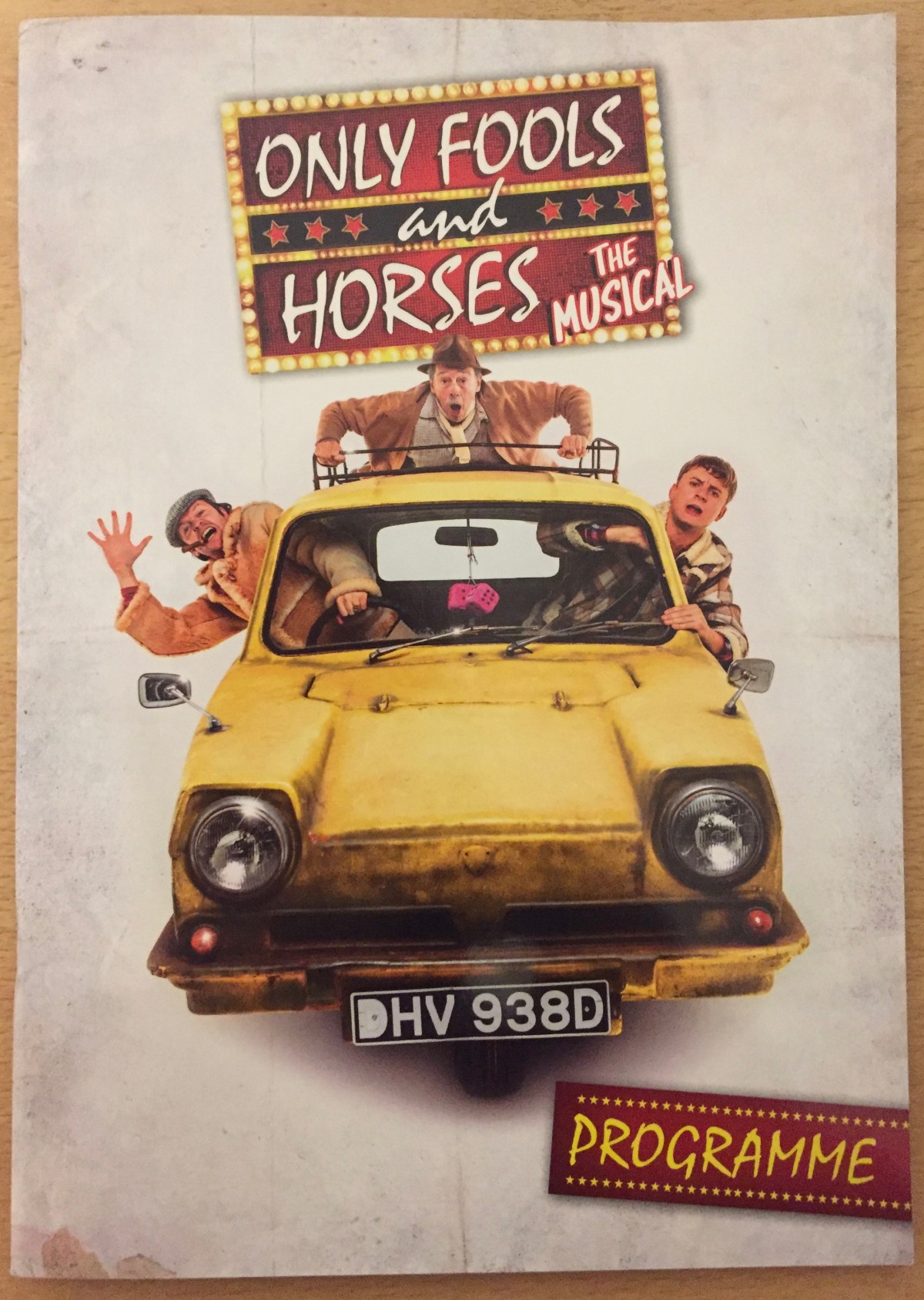 Cover of the programme for Only Fools And Horses, The Musical. Below the show logo is a large image of the yellow three-wheeled Reliant Regal van facing us, with 2 pink fluffy dice hanging from the rear view mirror in the centre. Del Boy and Rodney are leaning out of their side windows, with Del Boy waving and smiling with a cigar in his mouth, while Grandad is on the roof rack holding on tightly.