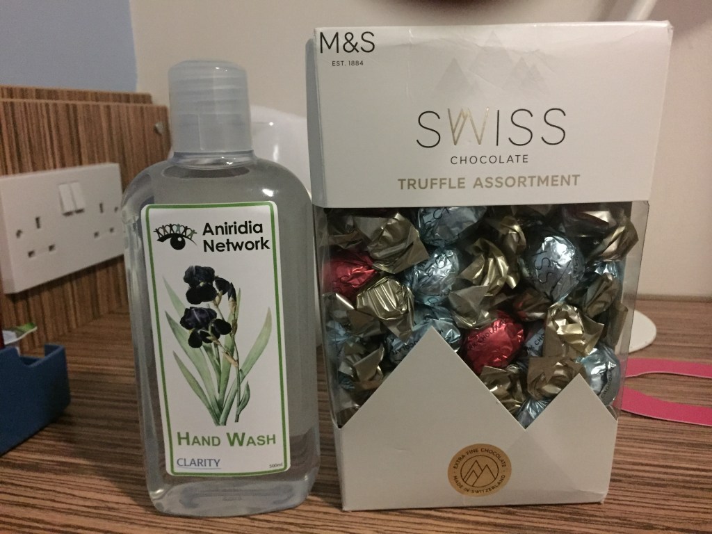 A tall bottle of Hand Wash, with he Aniridia Network logo and a black iris flower on the label, next to a big box of M&S Swiss Chocolate Truffles.