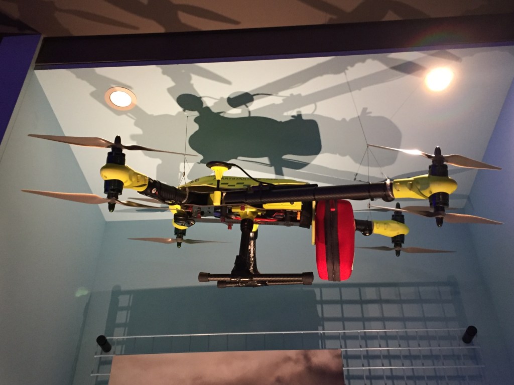 A black and yellow drone machine, with legs sticking out of each of the 4 corners of the central section, and a small propeller on the end of each of those legs.