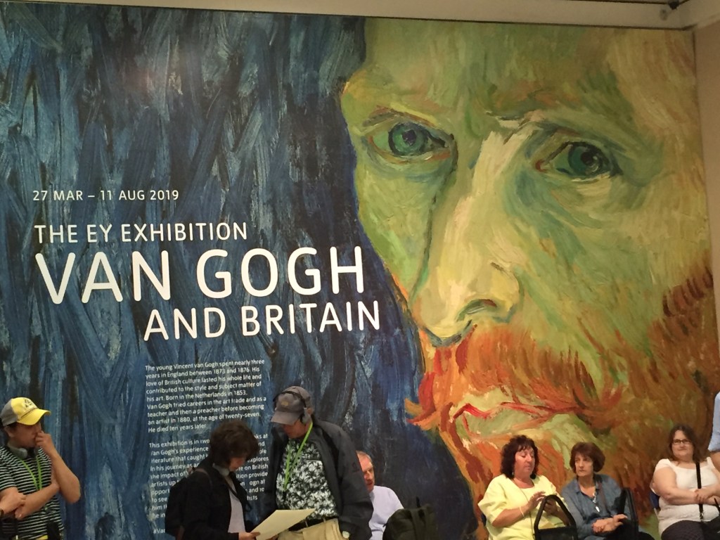 A large portrait of Vincent Van Gogh's face on a wall in Tate Britain. Text next to it says 27 March to 11 August 2019, The E.Y. Exhibition, Van Gogh And Britain. Below that is a description of the exhibition in smaller text