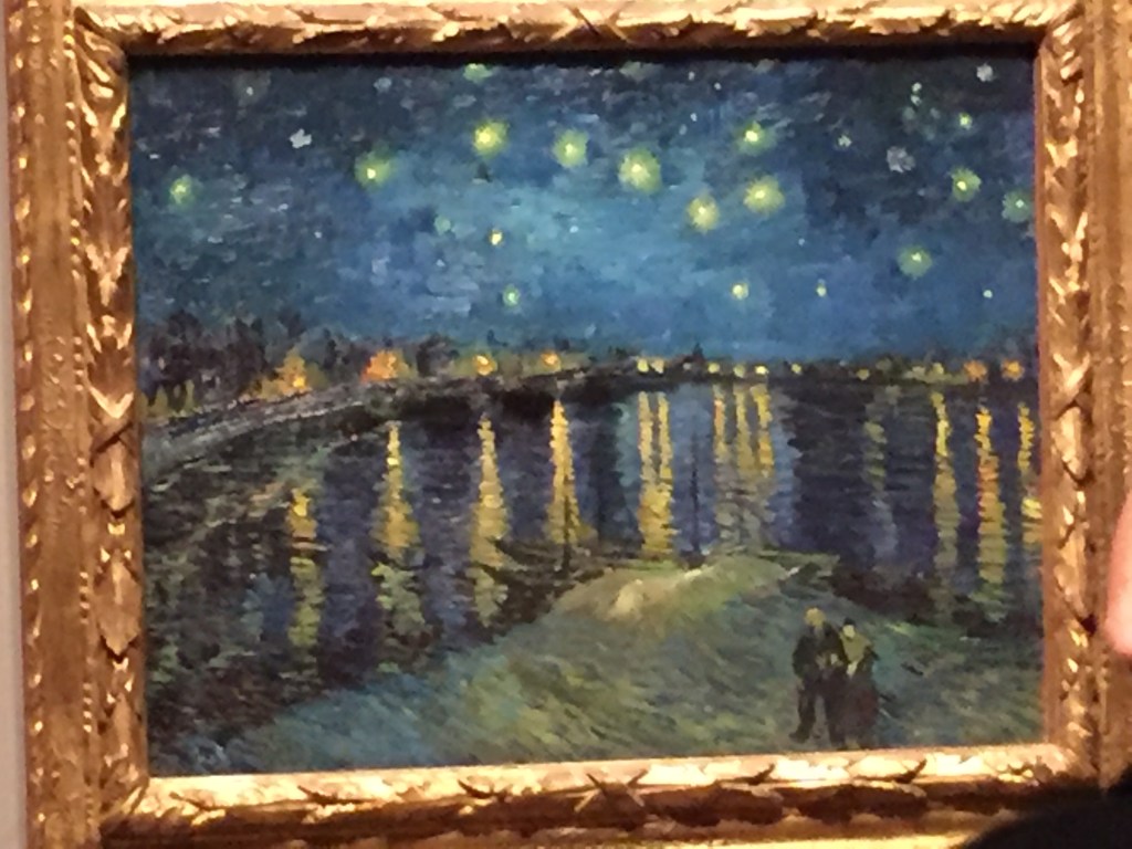 Van Gogh painting of a wide river stretching into the distance, with lights from buildings along each riverbank, and stars shining down from the sky. At the bottom of the painting, 2 people stand together on a patch of land overlooking the river.