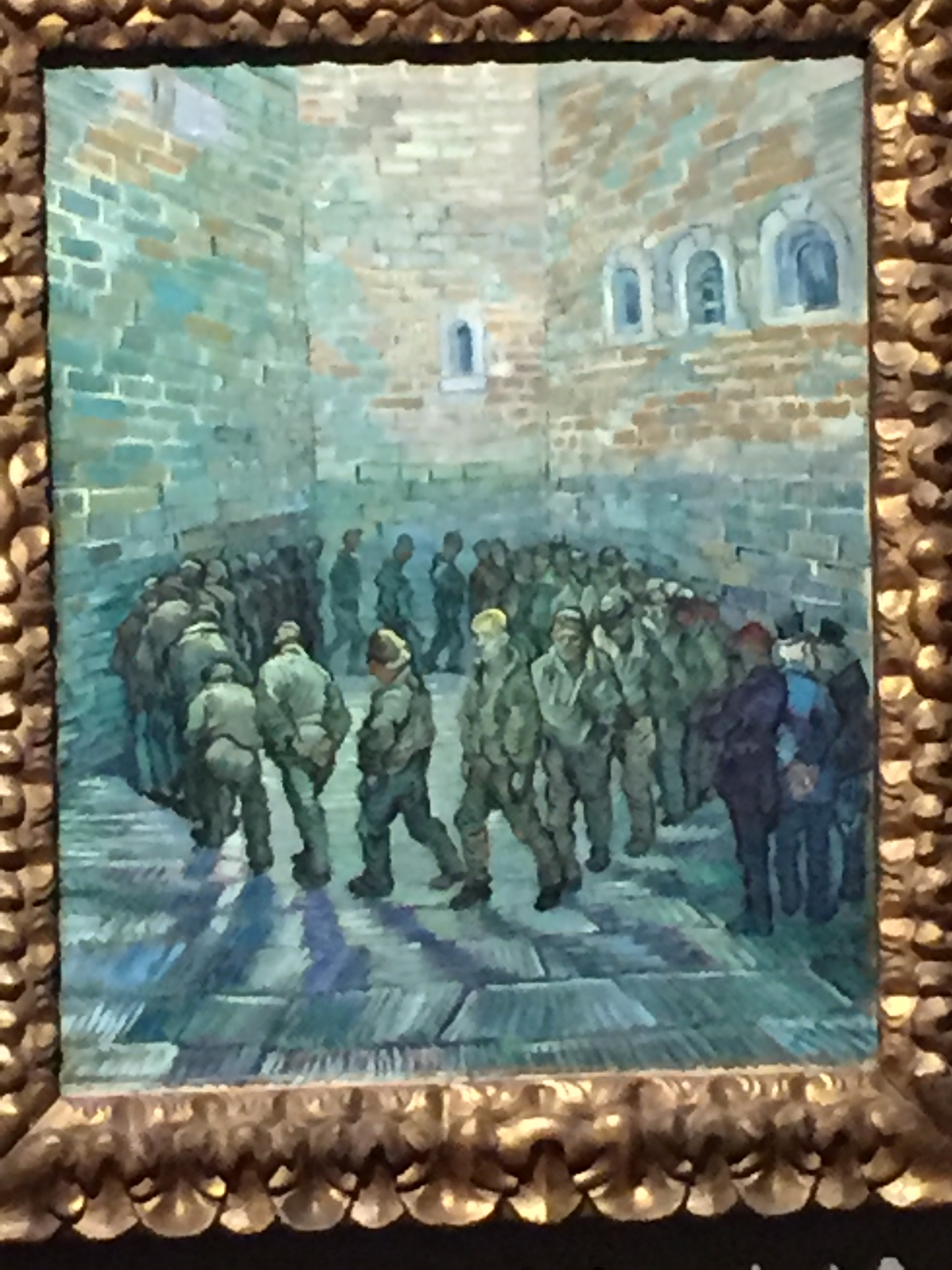 Van Gogh painting showing a group of men in plain grey uniforms, trudging around in a circle in a small courtyard, with high brick walls all around them, as 3 people watch from the bottom-right corner.