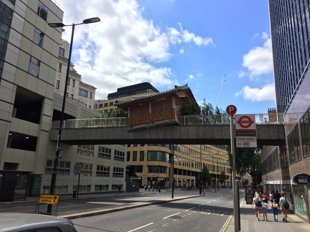 A model of a small bungalow resting on a footbridge across the road, its uneven angle suggesting that it has fallen from the sky and landed there.