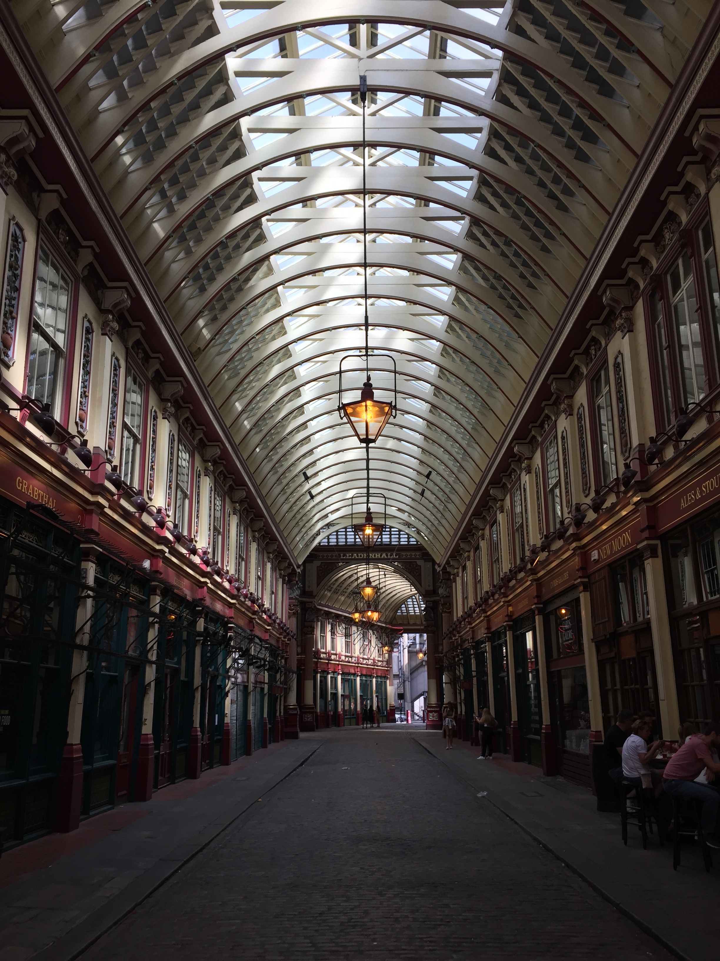 Interior of the historic Leadenhall Market, with many shops and restaurants lining each side of the street. The street is covered by a curved roof, with windows all along its central length that allow daylight in.