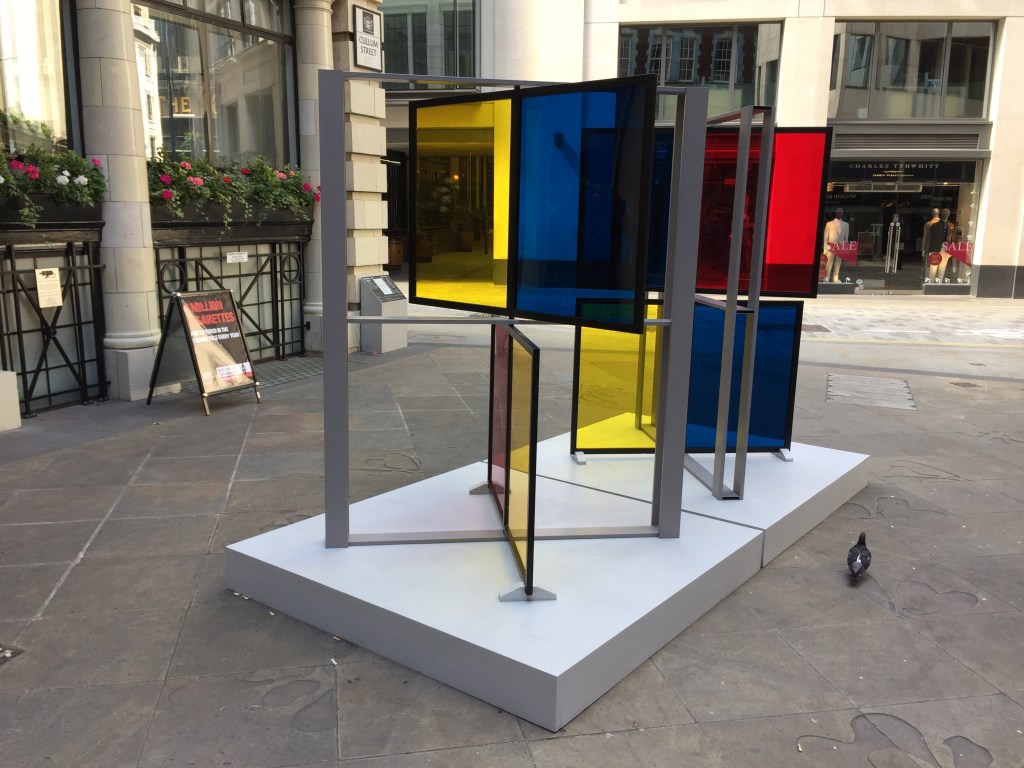 Sculpture made of 8 large square panes of glass, each a different colour, being either yellow, red or blue. They are arranged in 2 large metal grids, which stand diagonally on 2 adjacent white squares, using opposite diagonals. In each grid the top and bottom pair of panes each swivel in the centre, and have been turned to face in different directions.