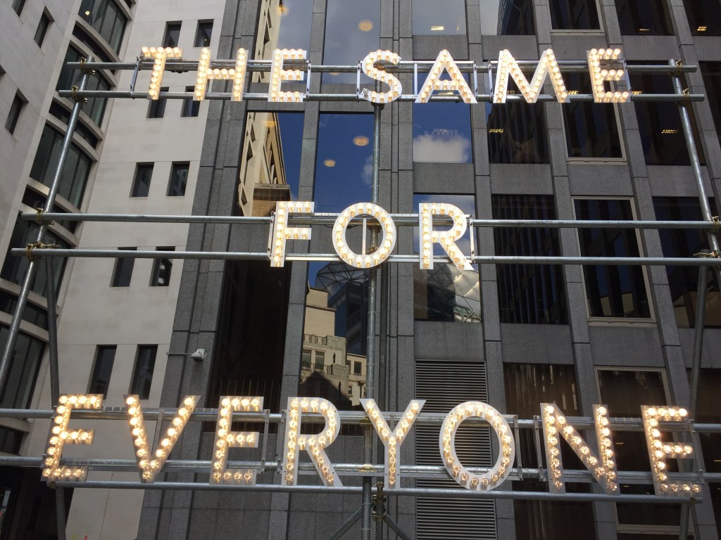 A tall, large metal stand, on which there are large letters made up of multiple small lightbulbs, spelling out the words The Same For Everyone.