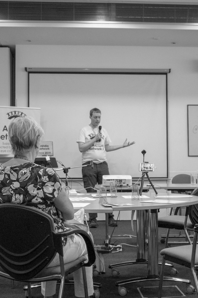 A black and white photo of Glen standing and talking into a microphone. He is wearing a white t-shirt with the Aniridia Network logo on it, while next to him is a tall free-standing banner for Aniridia Network UK, and a laptop on a podium with an Aniridia Network Conference 2019 sign on its open lid.