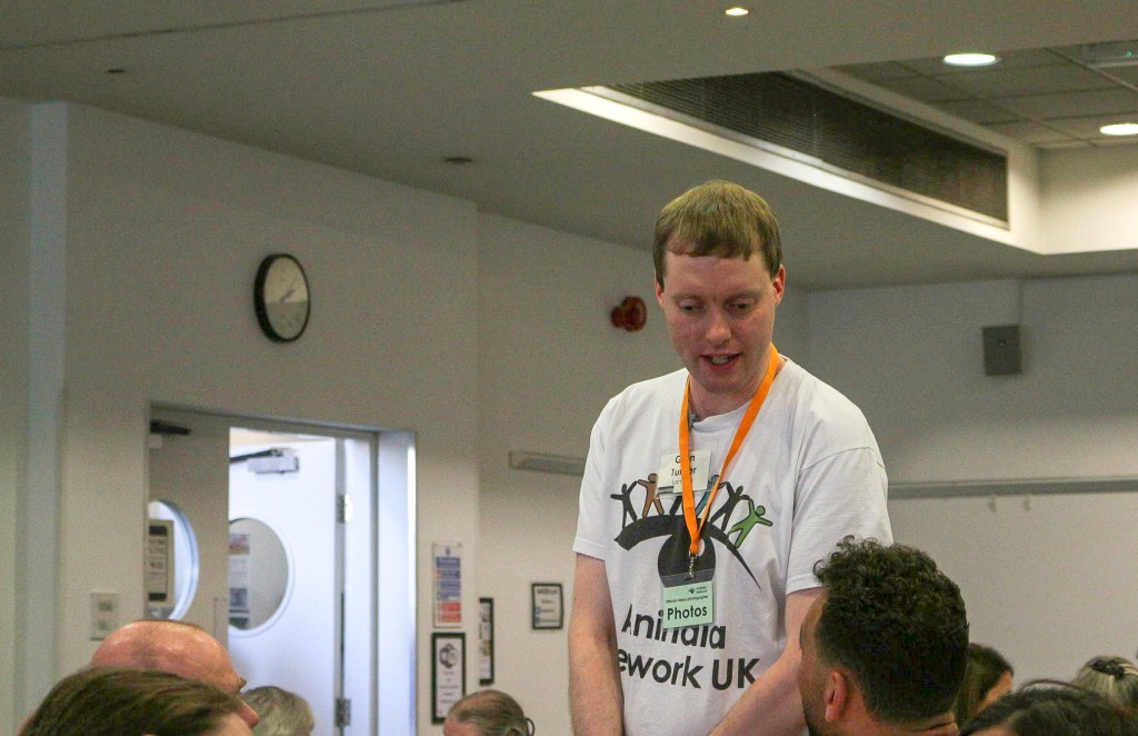 Glen talking to other delegates at the Aniridia Network Conference. He is wearing a white t-shirt with the Aniridia Network logo on, which features a line of differently coloured gingerbread-men-type figures standing on the eyebrow of an eye, with a black pupil beneath. Around his neck, Glen also has an orange lanyard, on the bottom of which is a plastic sleeve with a card saying Photos, designating him as an official photographer for the event.