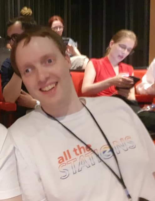 Glen smiling while sitting in the audience for An Evening Of Unnecessary Detail. He is wearing a white t-shirt with the multi-coloured logo for All The Stations on it, and has his monocular telescope on a lanyard around his neck.