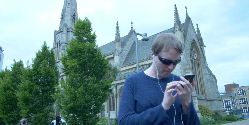 Standing in front of a large church, Glen is wearing earphones and sunglasses while looking at his phone.