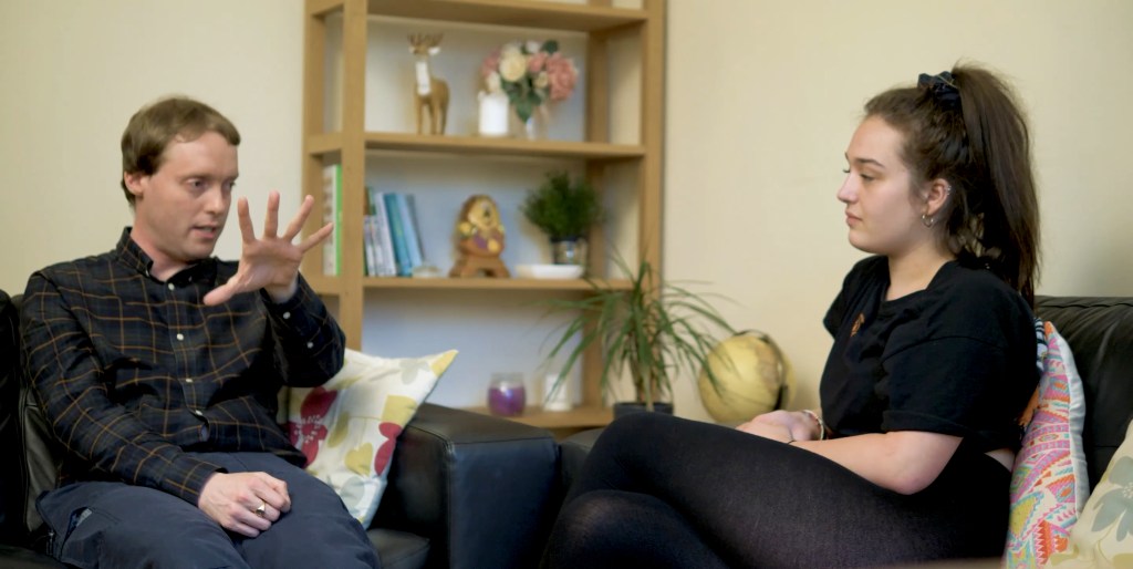 Glen gesturing with his hand out in front of him as he is interviewed by Stella in a living room.