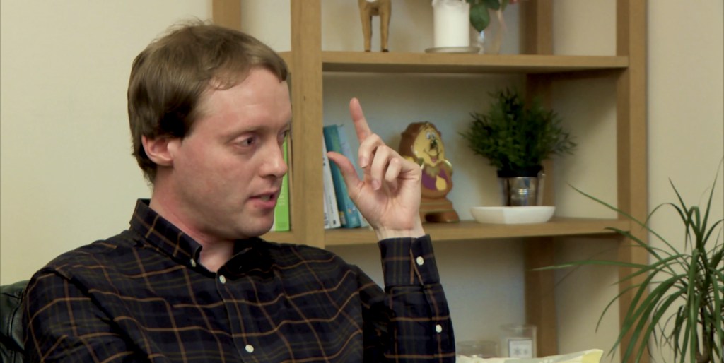 Close-up of Glen gesturing with his fingers near his eye, during his interview in a living room.