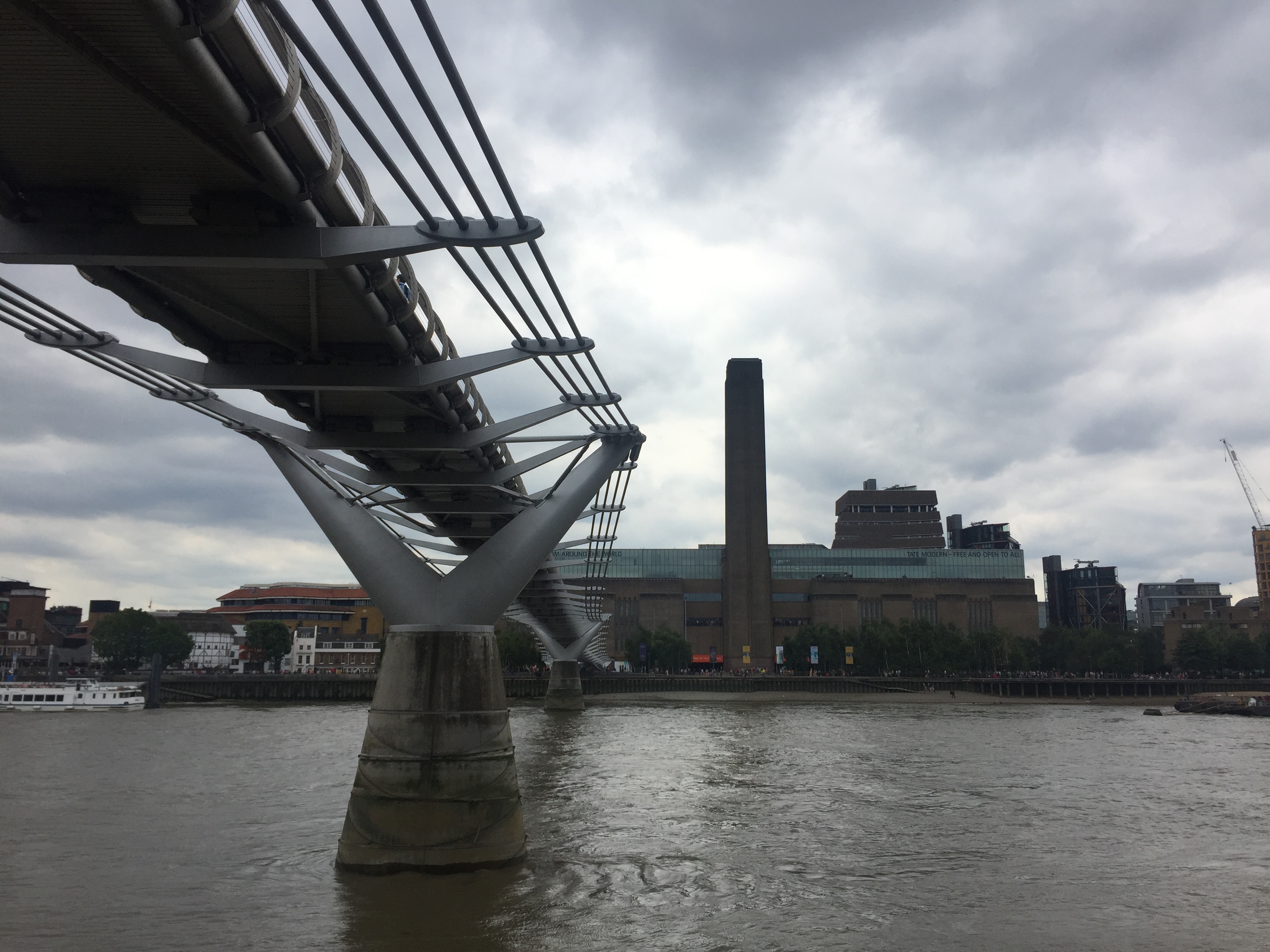 View from the North Bank of the River Thames towards the Tate Modern on the opposite bank, with its tall chimney sticking far up above the rest of the building. The Millennium Bridge spans the river to the left, and is above us, so we can see its underside.