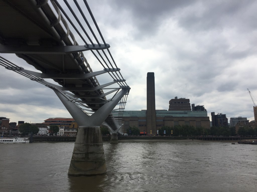View from the North Bank of the River Thames towards the Tate Modern on the opposite bank, with its tall chimney sticking far up above the rest of the building. The Millennium Bridge spans the river to the left, and is above us, so we can see its underside.