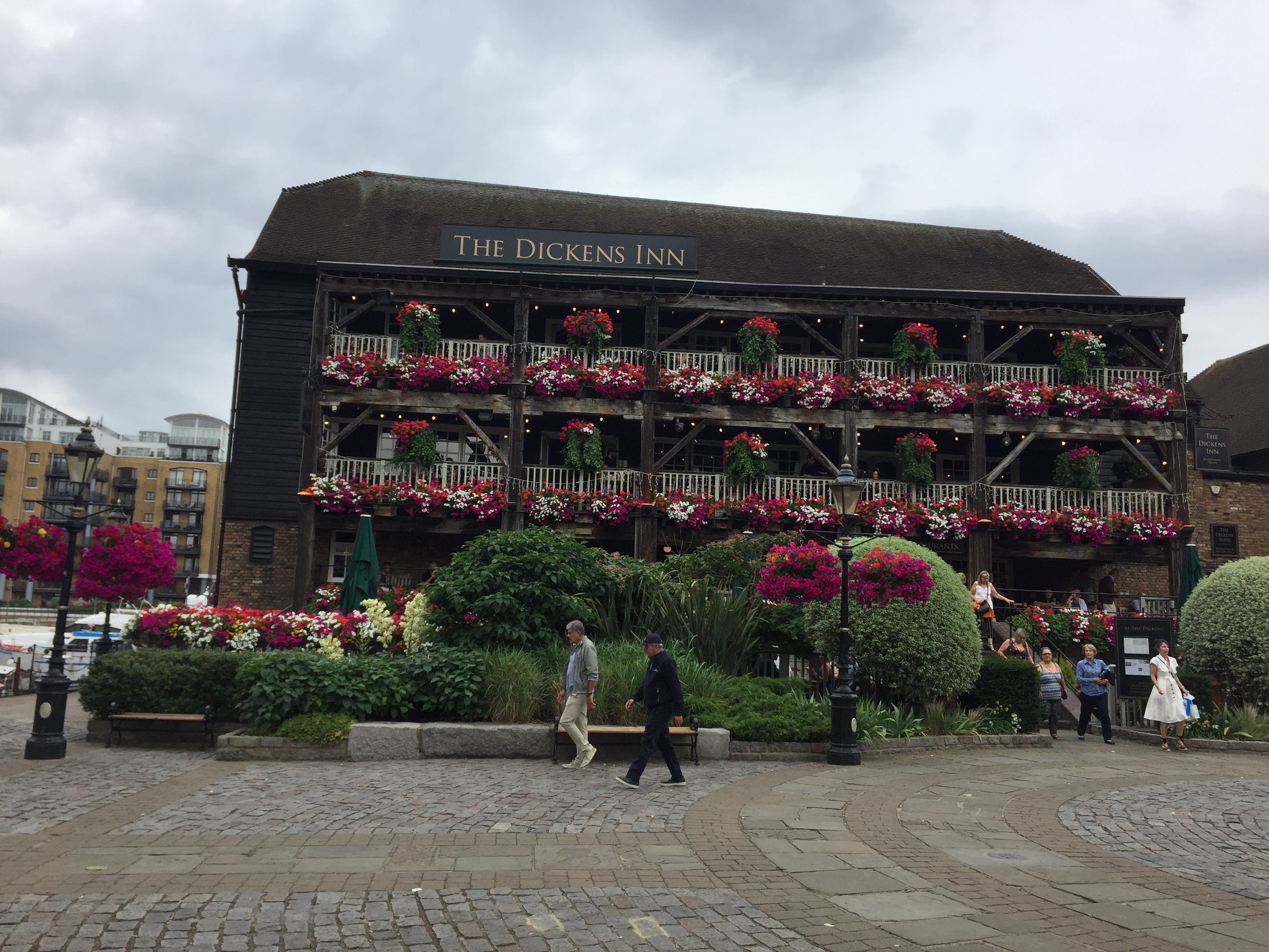 The Dickens Inn, a large pub with 3 storeys. The upper 2 levels have balconies with colourful flowers decorating the fencing. Green bushes and more colourful bunches of flowers surround the seating area outside the building, obscuring it from view, including a couple of lampposts with hanging baskets attached, containing even more flowers.