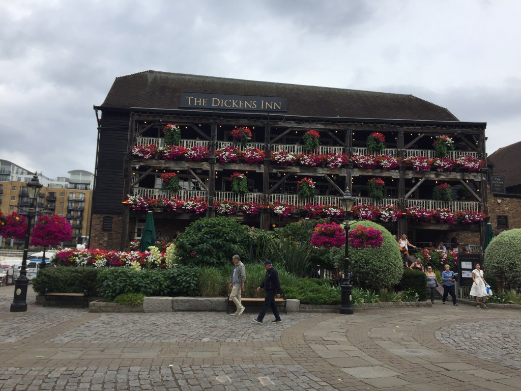 The Dickens Inn, a large pub with 3 storeys. The upper 2 levels have balconies with colourful flowers decorating the fencing. Green bushes and more colourful bunches of flowers surround the seating area outside the building, obscuring it from view, including a couple of lampposts with hanging baskets attached, containing even more flowers.