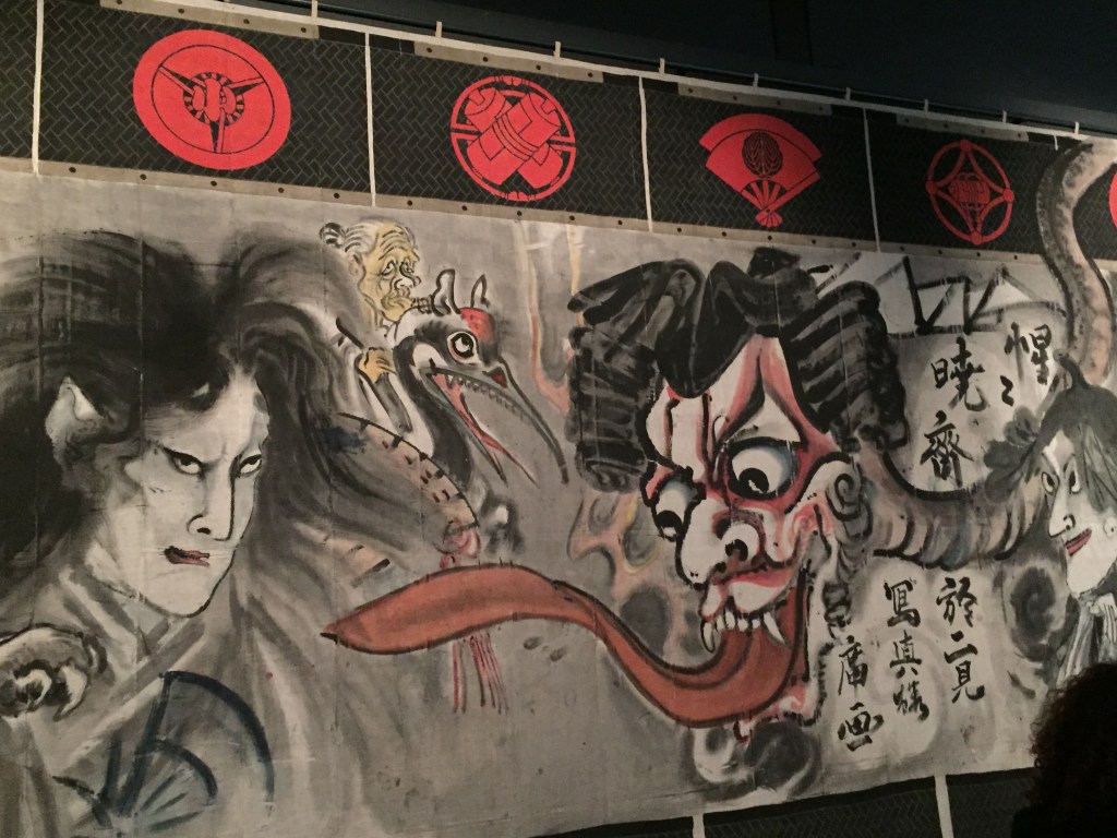 Section from a large manga wall mural. On the right is a large head with a long red tongue snaking out of its mouth towards a man on the left side. In the centre at the back is a large bird with teeth along the inside of its beak, with an elderly character riding on its back.