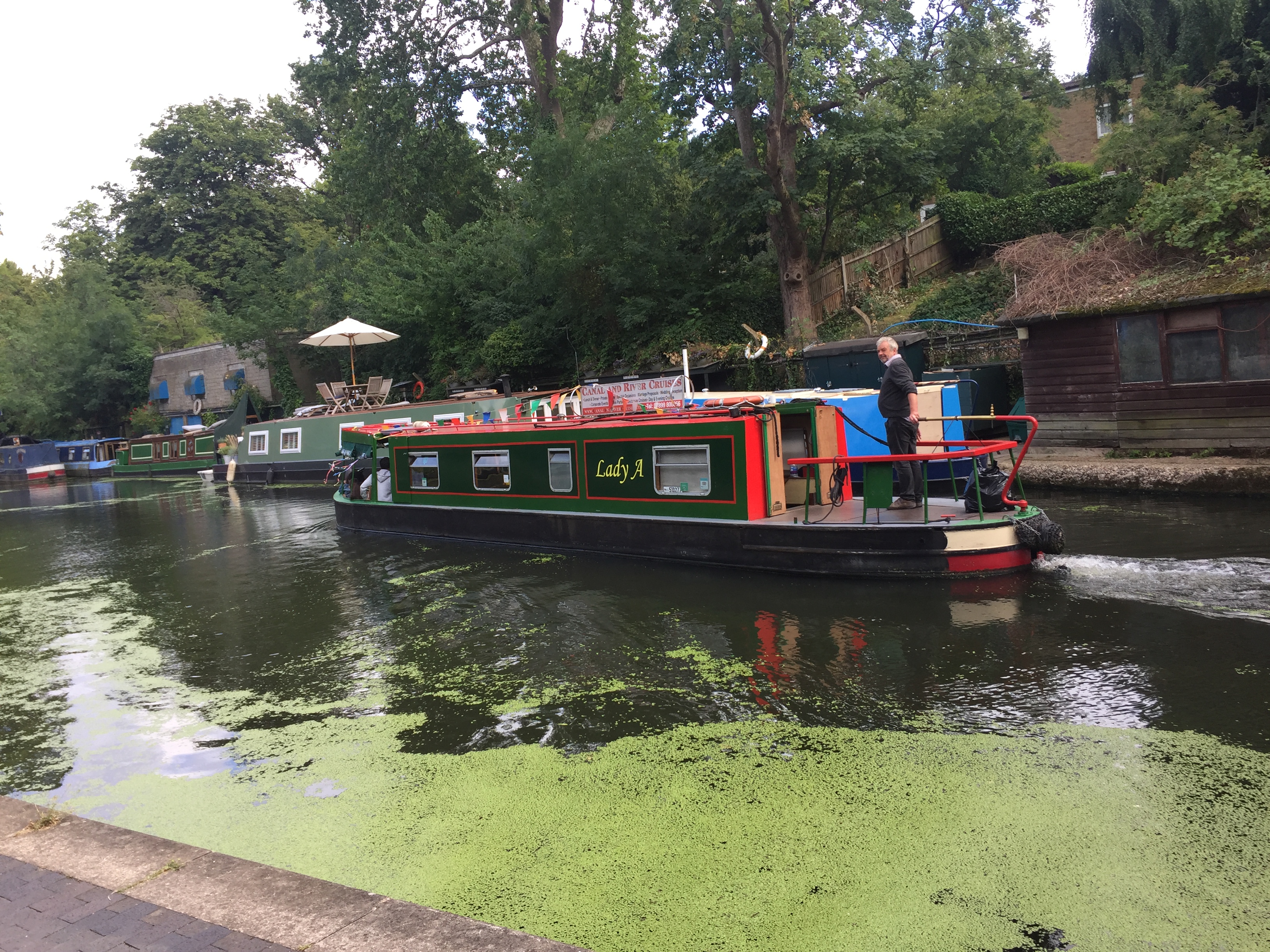 A man sailing a long barge called Lady A down Regent's Canal, passing other barges moored up to the side.