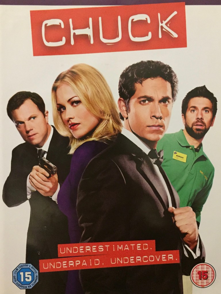 Image of John, Sarah, Chuck and Morgan from the series Chuck. Sarah and Chuck are at the front, standing back to back as they look sideways towards us, with Sarah in a purple dress and Chuck in a black suit and bow tie. To their left, John is in a suit pointing a gun, while to their right Morgan is leaning out from behind them in his green Buy More uniform to see what's going on.