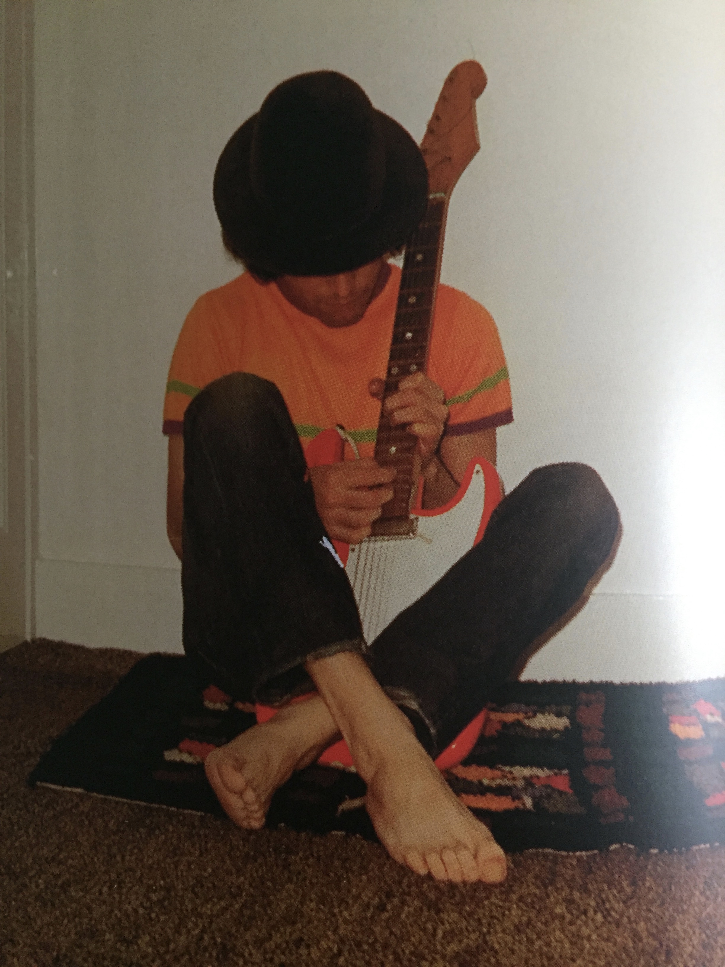 Photo of a young Freddie Mercury, wearing an orange t-shirt and black trousers and nothing on his feet, sitting cross-legged on the floor as he plays the guitar.