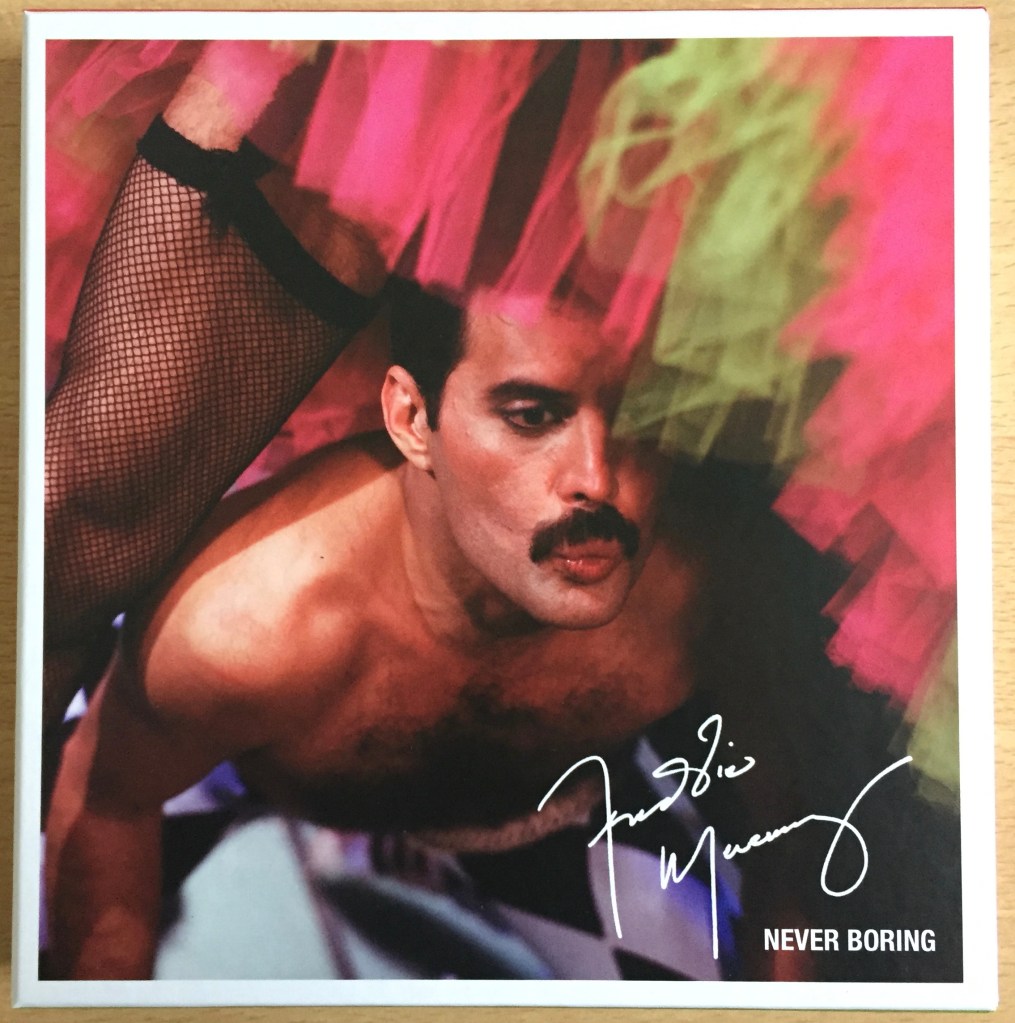 Cover of the Never Boring box set by Freddie Mercury. The image shows Freddie, topless and pouting his lips, as he dances between another person's legs, which are clad in fishnet stockings beneath a green and pink skirt. Freddie's signature is in white in the bottom right corner of the cover, above the title Never Boring.