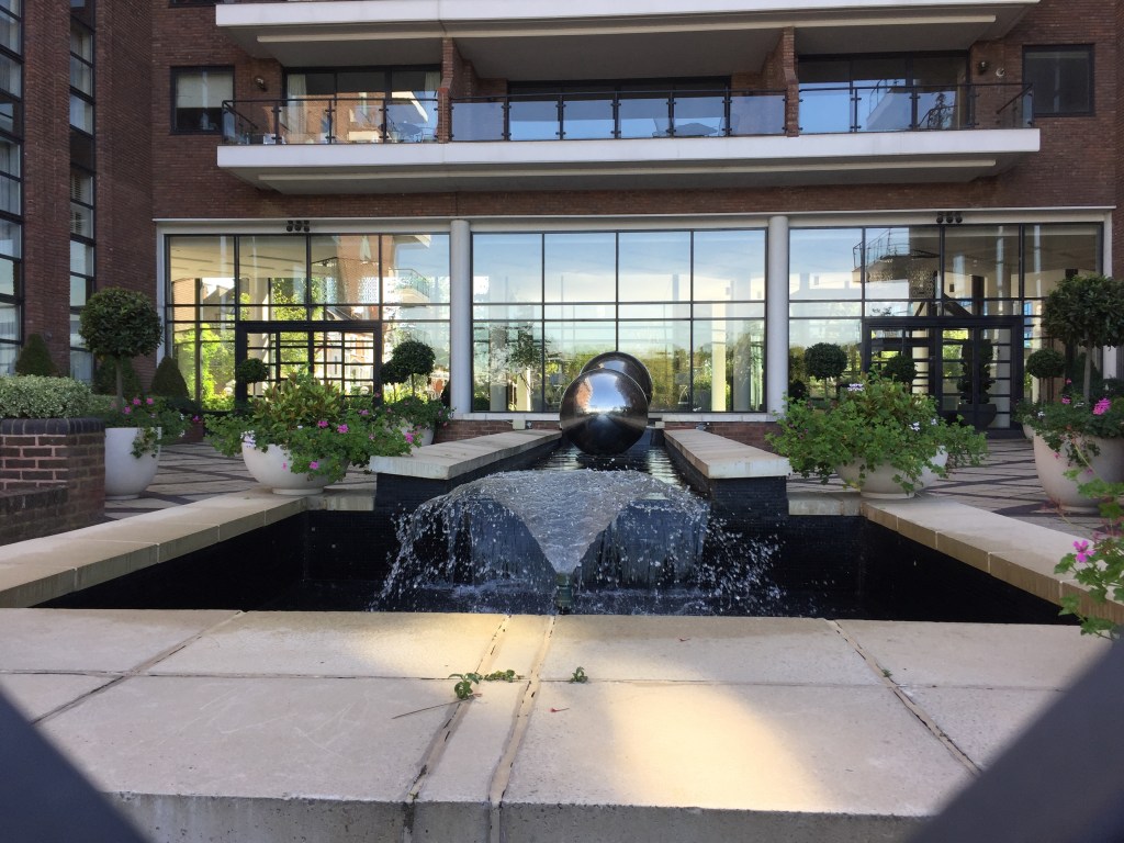 Large water feature, consisting of a long narrow trough of water, the end of which cascades the water into a wide rectangular pool lower down. The long trough also contains a couple of large shiny silver balls, and there are large pots containing flowers on the pavement next to the trough wall on each side.