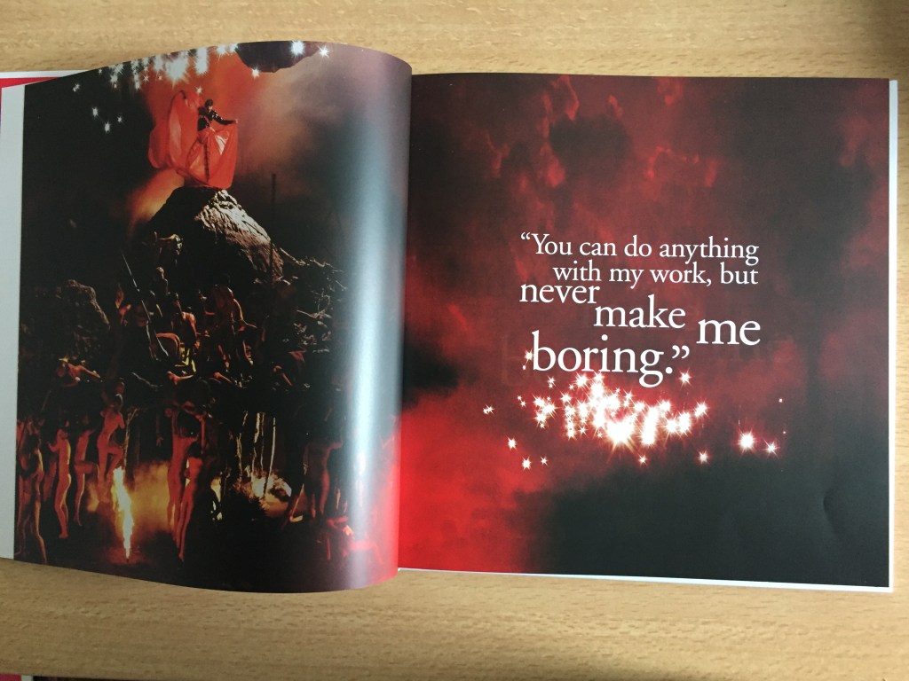 A 2-page spread from the Never Boring book about Freddie Mercury. The left page shows Freddie fanning out a red cape behind him as he stands on a tall model of a mountain or volcano, while people gather around the bottom as if trying to reach him and worship him. The right page has the quote You can do anything with my work, but never make me boring.