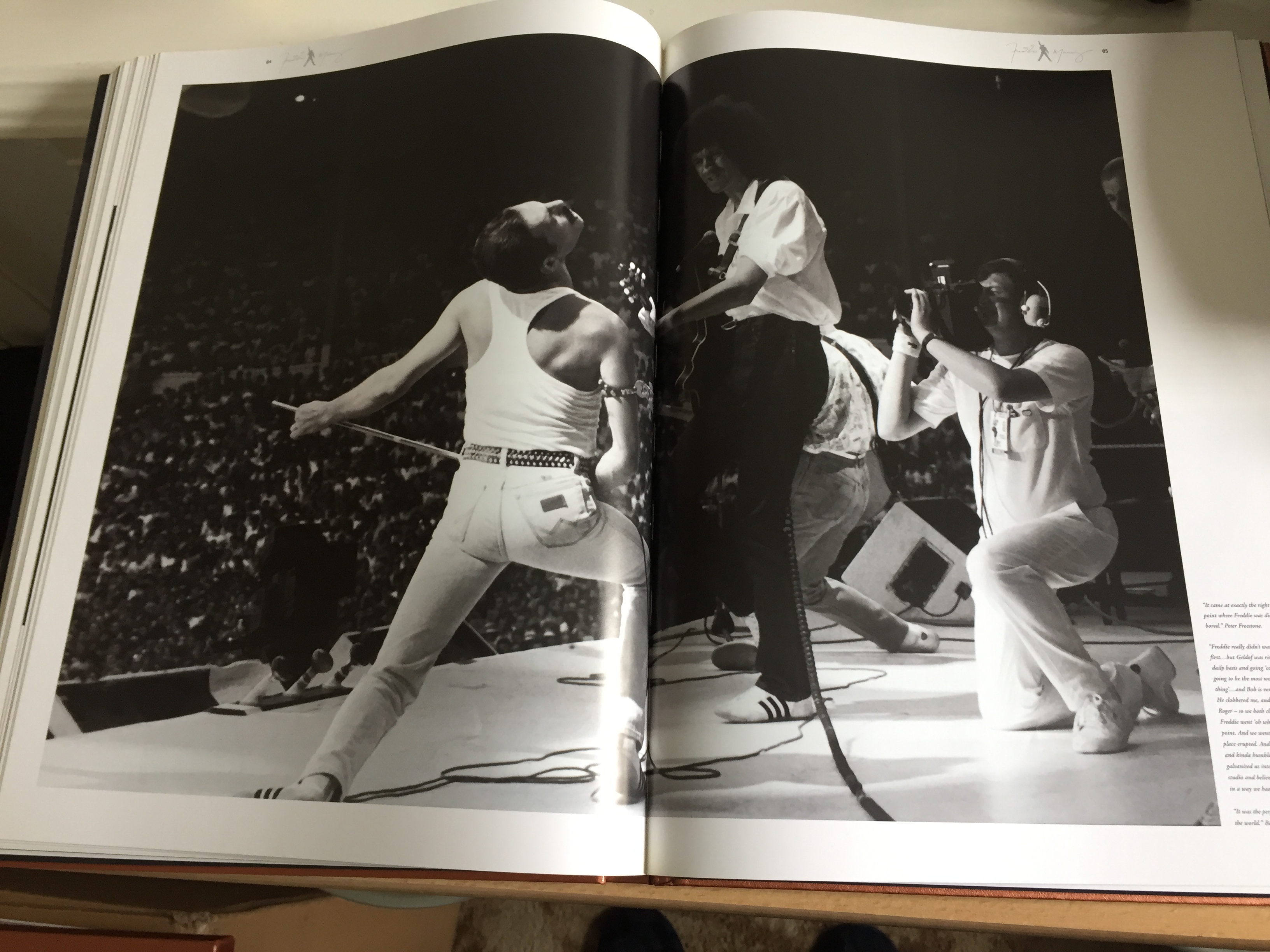 A double-page spread from the Freddie Mercury Solo Collection book, across which is a black and white photo of Freddie Mercury and Brian May performing together on stage, with a cameraman kneeling behind them as he films them close-up.