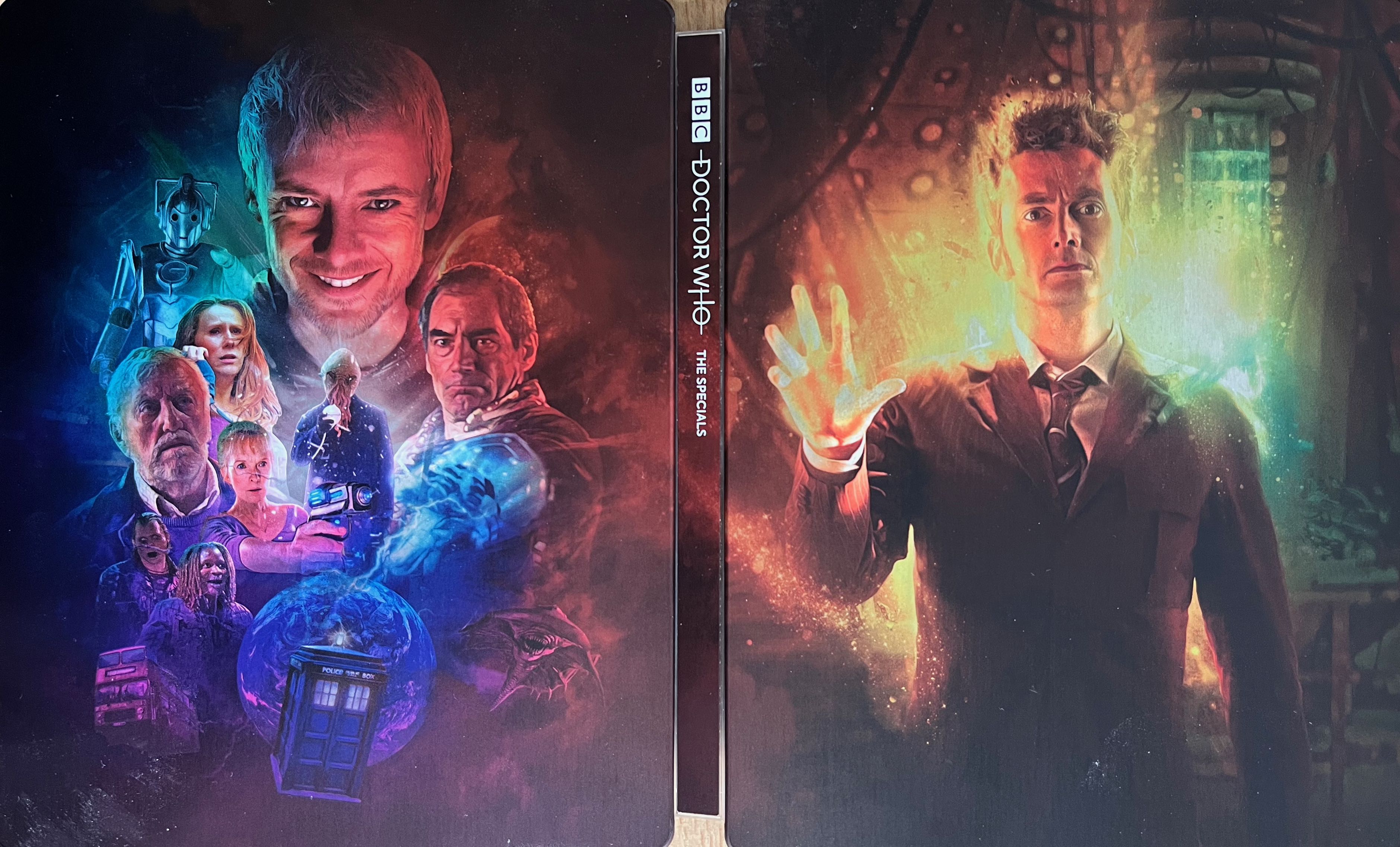 The full cover spread artwork for the Doctor Who 2008 to 2010 Specials Blu-ray steelbook. The front cover shows David Tennant as the Doctor in a brown suit, standing in the Tardis, with glowing yellow regeneration energy around him.
Meanwhile the back cover shows the main enemies and friends that he encounters during the episodes. There is a large picture of John Simm's grinning face as the Master at the top, with figures below including Donna, her grandfather Wilfred, Timothy Dalton as a Time Lord, and an Ood with spaghetti-like strands hanging from its mouth, while the Tardis is at the very bottom of the cover.