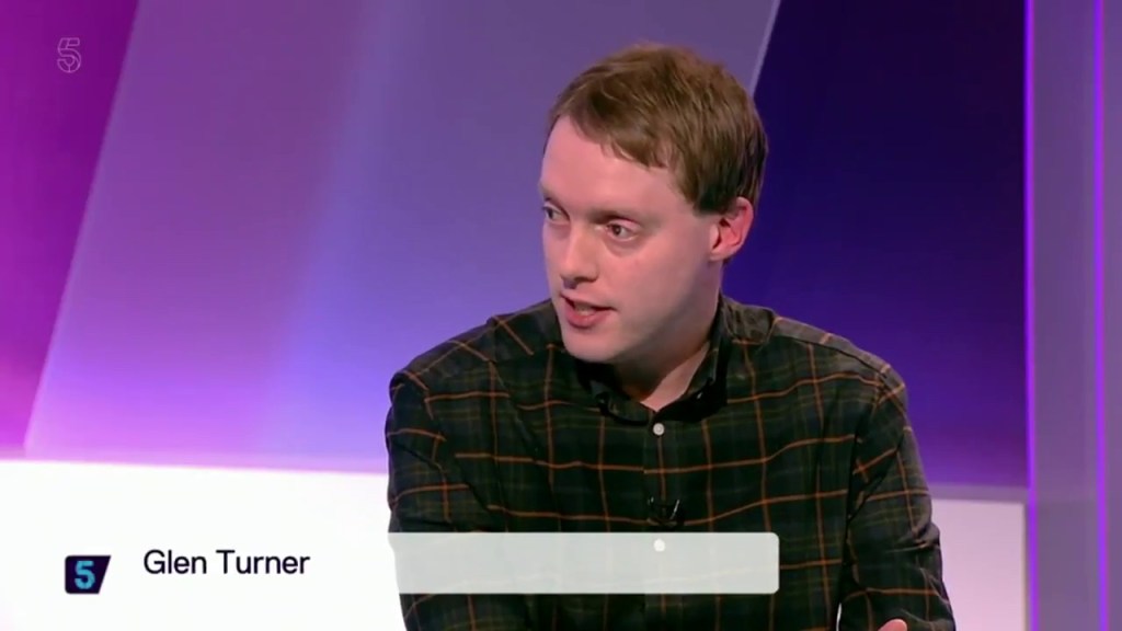 Close-up of Glen on 5 News, wearing a green chequered shirt with the studio's purple backdrop behind him. His name is shown on a caption at the bottom of the screen.