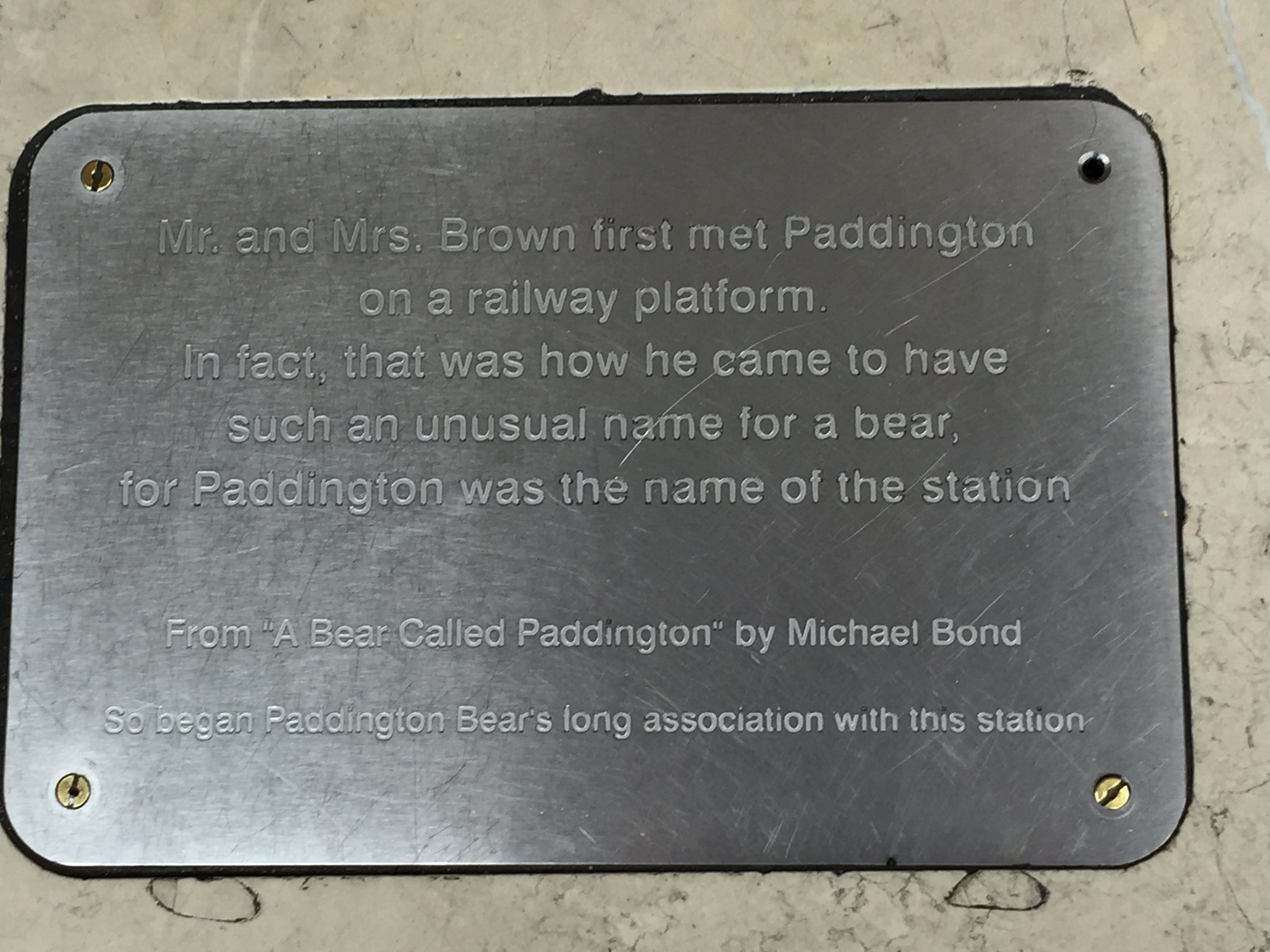 Silver plaque by the Paddington Bear statue at Paddington Station. The text says Mr and Mrs Brown first met Paddington on a railway platform. In fact, that was how he came to have such an unusual name for a bear., for Paddington was the name of the station. From A Bear Called Paddington by Michael Bond. So began Paddington Bear's long association with this station.