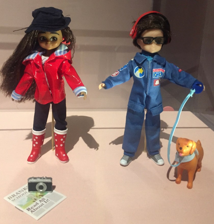 2 dolls, one of which is wearing dark glasses and an all-in-one blue suit, and has a golden coloured dog on a lead. The other doll is wearing a shiny red jacket, black trousers and red boots with white spots.