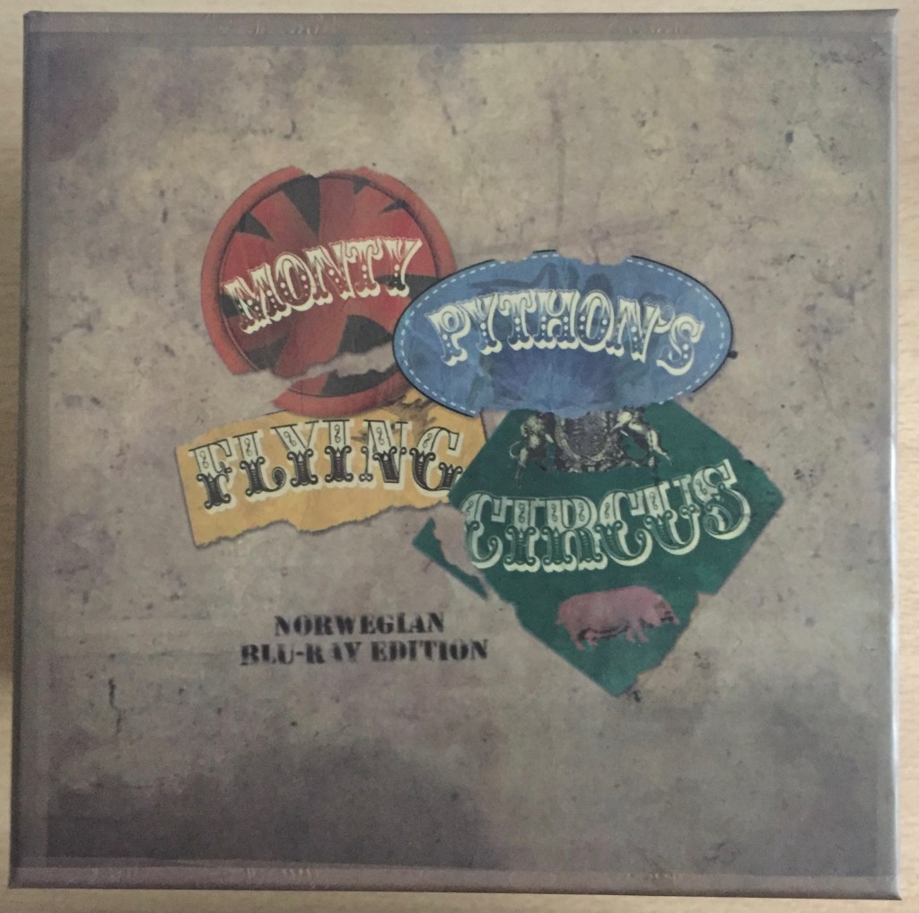 The lid of the cube shaped box set for Monty Python's Flying Circus. Random shapes in red, blue, yellow and green house each word of the show's title in turn, while text beneath says Norwegian Blu-Ray Edition. The background colour of the box has a very dull, dirty, worn appearance, as if the box is 50 years old like the programme itself.