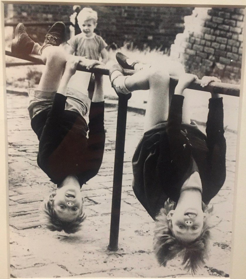 Black and white photo of 2 children hanging upside down from a metal railing outdoors, holding on to it with their hands and feet.