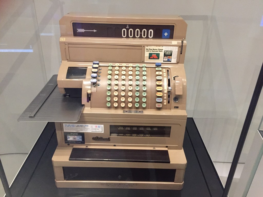 A large cash register till in a glass case at the Science Museum. It has many rows of buttons on the curved central section, a draw at bottom, and a narrow screen at the top with white numbers on a black background.