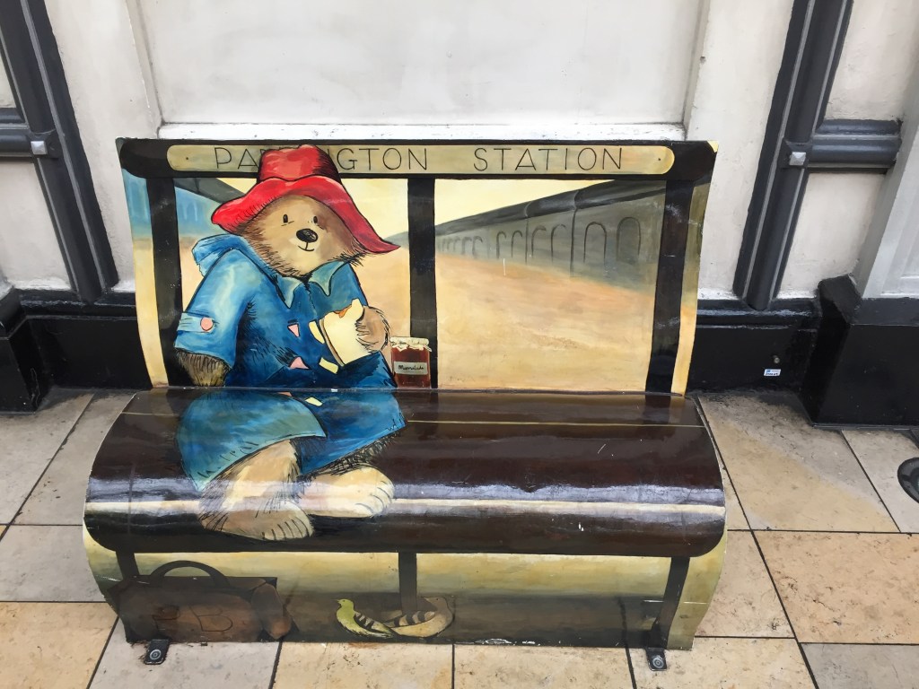 Bench at Paddington station, painted to look like Paddington Bear is sitting on it, in his red hat and blue duffel coat, smiling as he eats a sandwich, with his suitcase and a couple of pigeons on the floor in front of him.