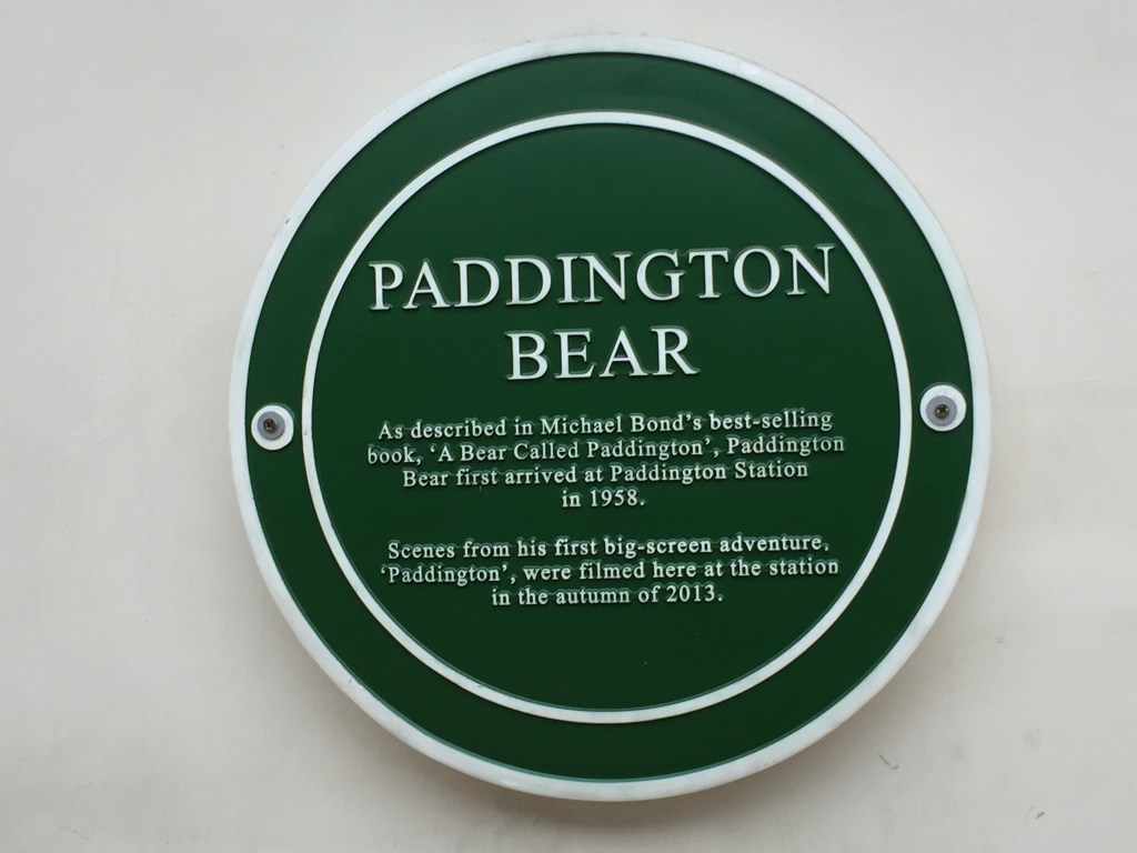 A round green plaque on the wall at Paddington station, dedicated to Paddington Bear. The text says as described in Michael Bond's best-selling book, A Bear Called Paddington, Paddington Bear first arrived at Paddington Station in 1958. Scenes from his first big-screen adventure, Paddington, were filmed here at the station in the autumn of 2013.