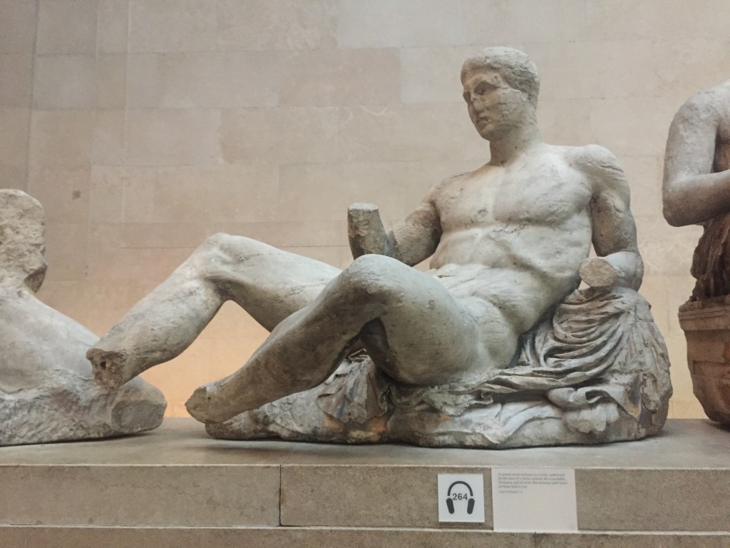 Statue of a naked young man reclining on a rock. The statue shows signs of wear and tear, and his hands and feet are missing.