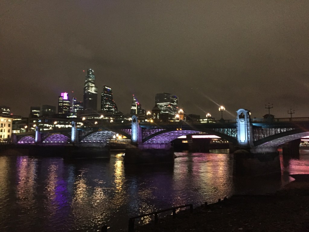 Southwark Bridge across the River Thames at night, with different colours of light illuminating the undersides of its ornate arches, including light shades of green, blue and purple.