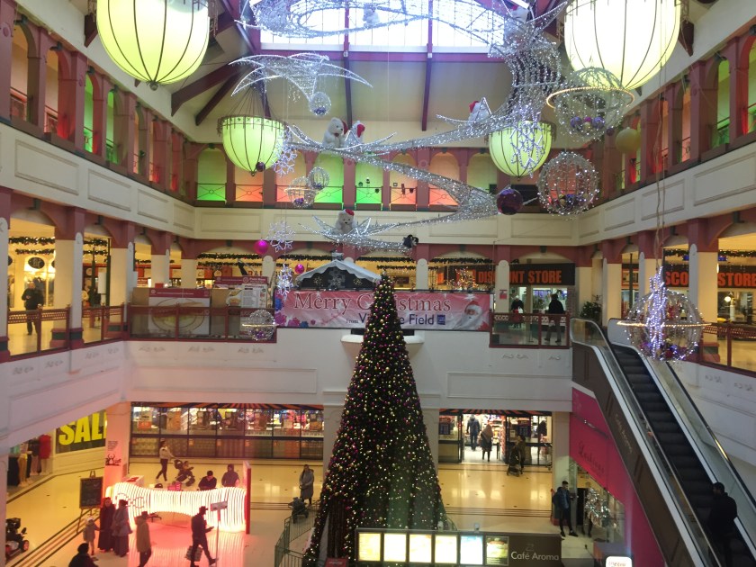 Interior of Vicarage Field Shopping Centre in Barking, on a first floor walkway that surrounds an area of the ground floor below. A large cone-shaped Christmas tree covered in lights on the ground floor stretches as high as he first floor. Above, large bulbous lights and many glittery decorations in different shapes hang from the ceiling.