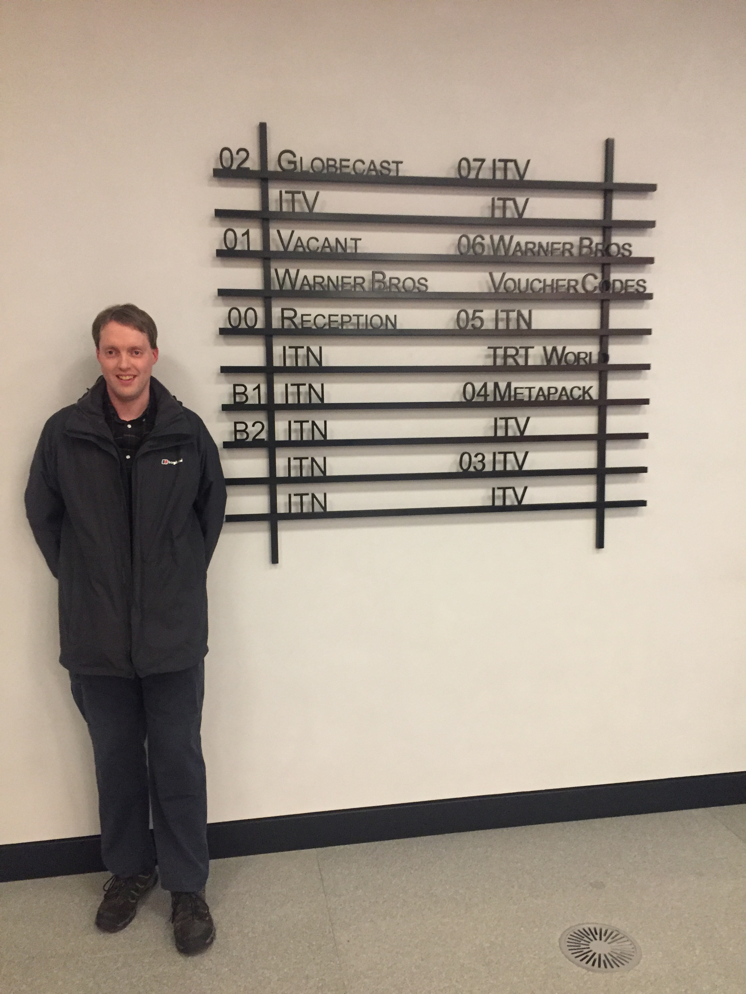 Glen smiling as he stands next to a tall sign, made of black bordered rows with black lettering, showing what's on each floor of the building. Most say ITN or ITV, but the sign also mentions Globecast, Warner Brothers, TRT World and Metapack, plus the reception desk.