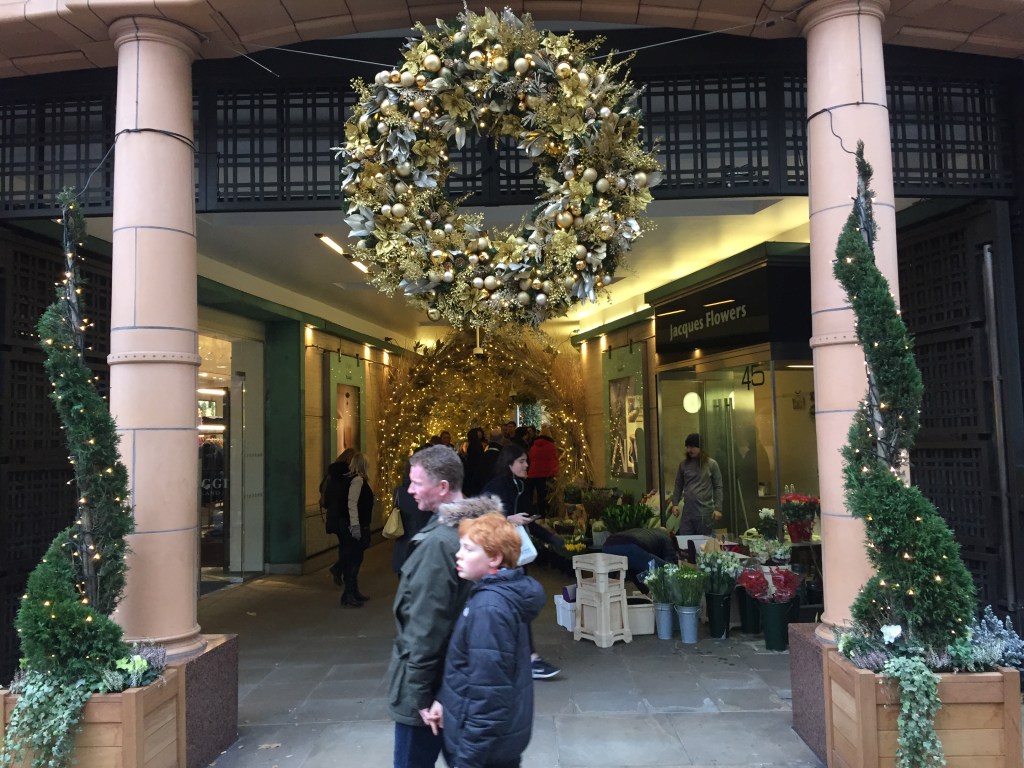 Entrance to Duke Of York Square shopping centre in Chelsea. A large wreath covered in golden baubles hangs over the entrance, while by a pillar on each side is a green tree that curves upwards in a snake-like way while covered in lights. Inside the corridor ahead is a tunnel made of yellow Christmas lights that people are walking through.