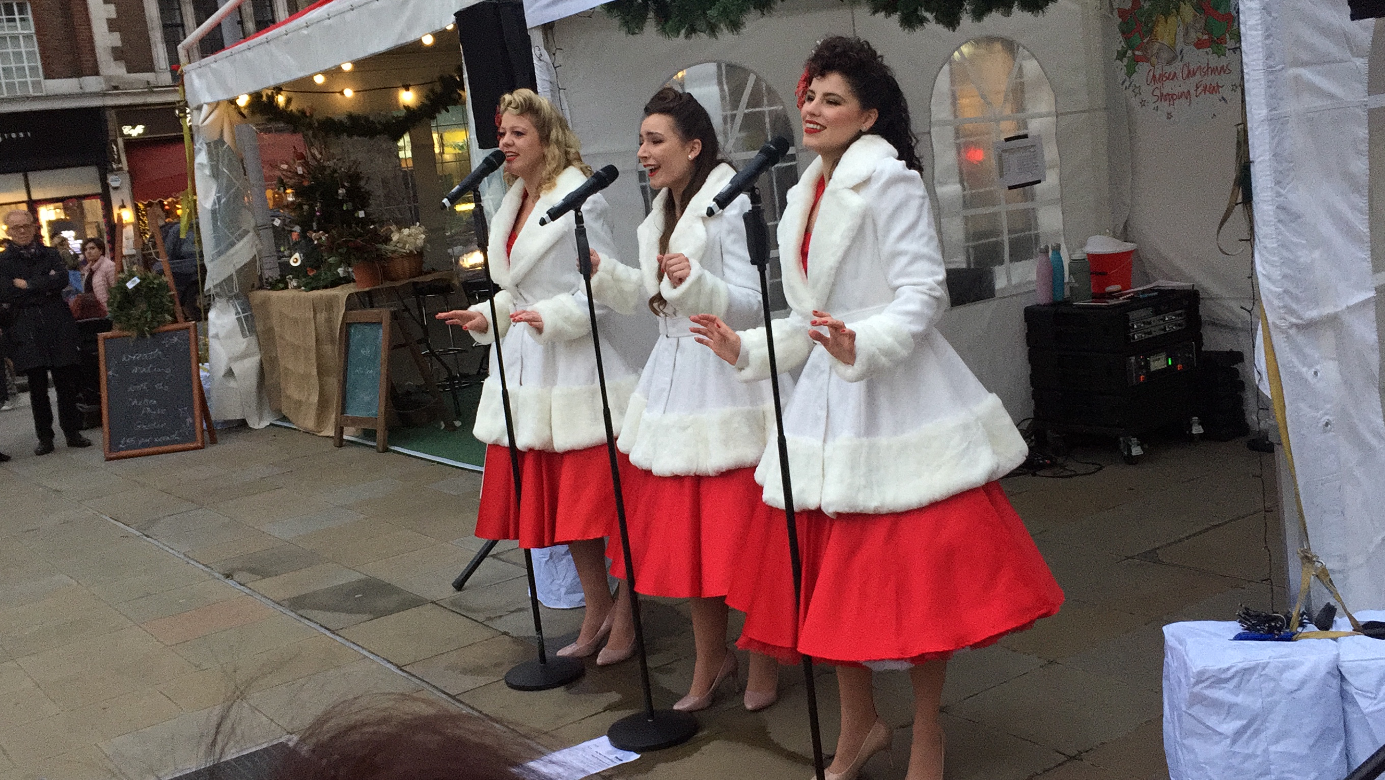 The Satin Dollz, 3 ladies smiling as they stand and sing at microphones, gesturing with their arms as they perform. Each of them is wearing a long white jacket with thick fluffy collars, cuffs and hem, over a large red skirt that swirls around them when they turn.