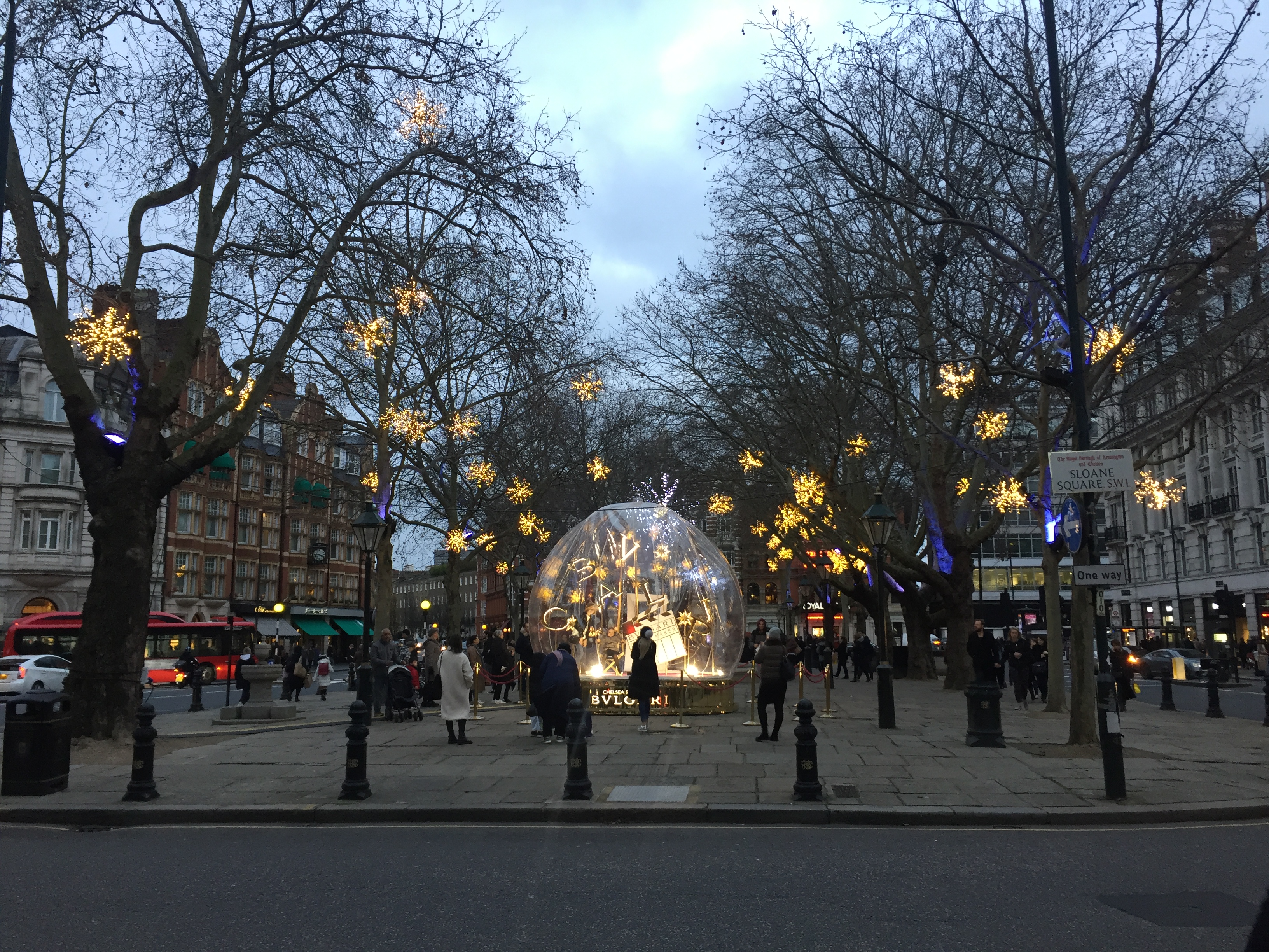 Sloane Square, with large lit-up star decorations hanging from trees along its length. At the end of the square nearest us is a large snow globe structure by a company called Bvlgar, in which people can sit on a director's chair to have their photos taken.