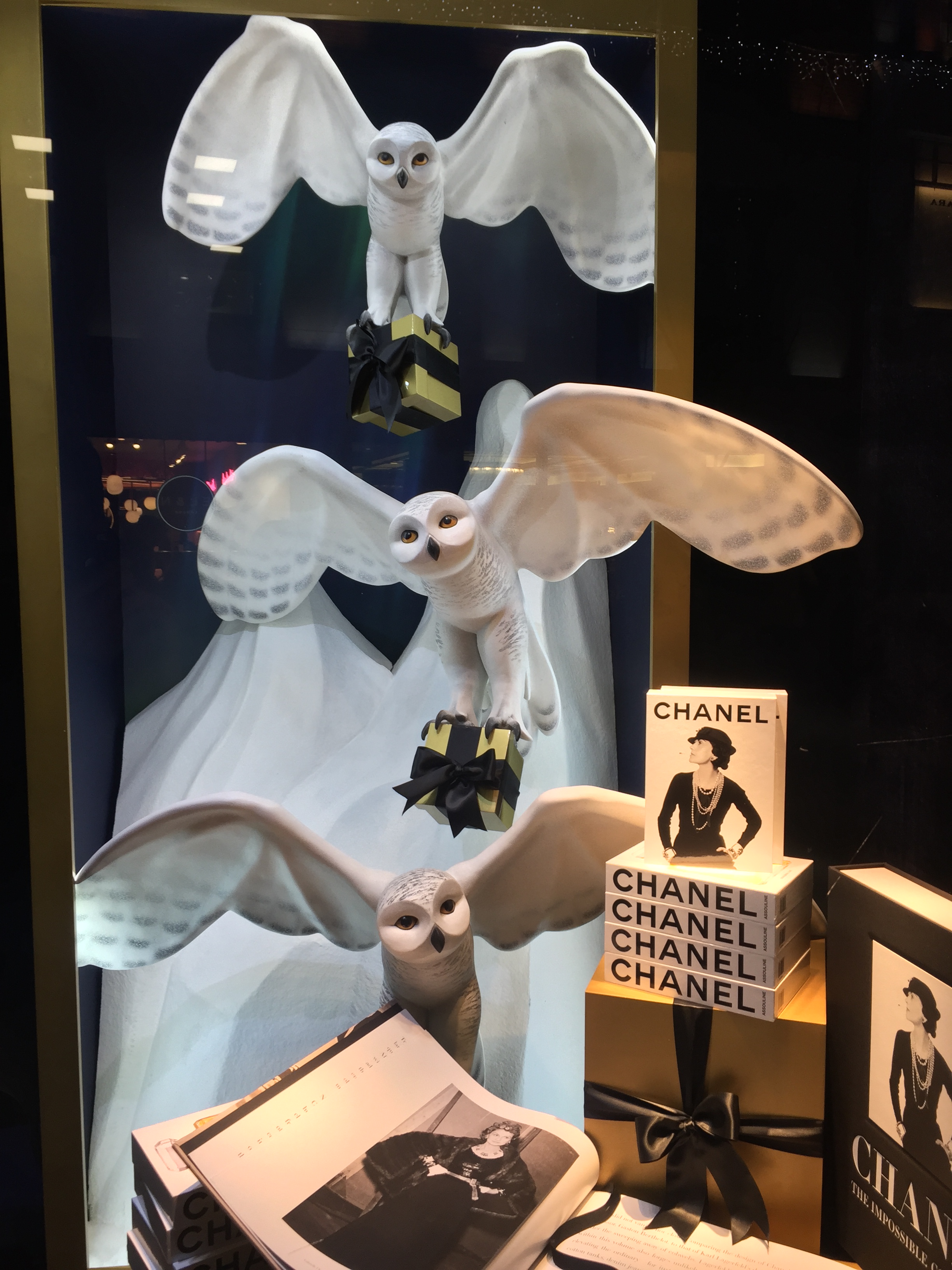 Models of 3 large white owls in flight, one above the other, in a Harrods window display. Each is carrying a small gift wrapped box with its talons. Beneath them is a pile of books about Chanel, with a lady posing with her hands on her hips on the cover.